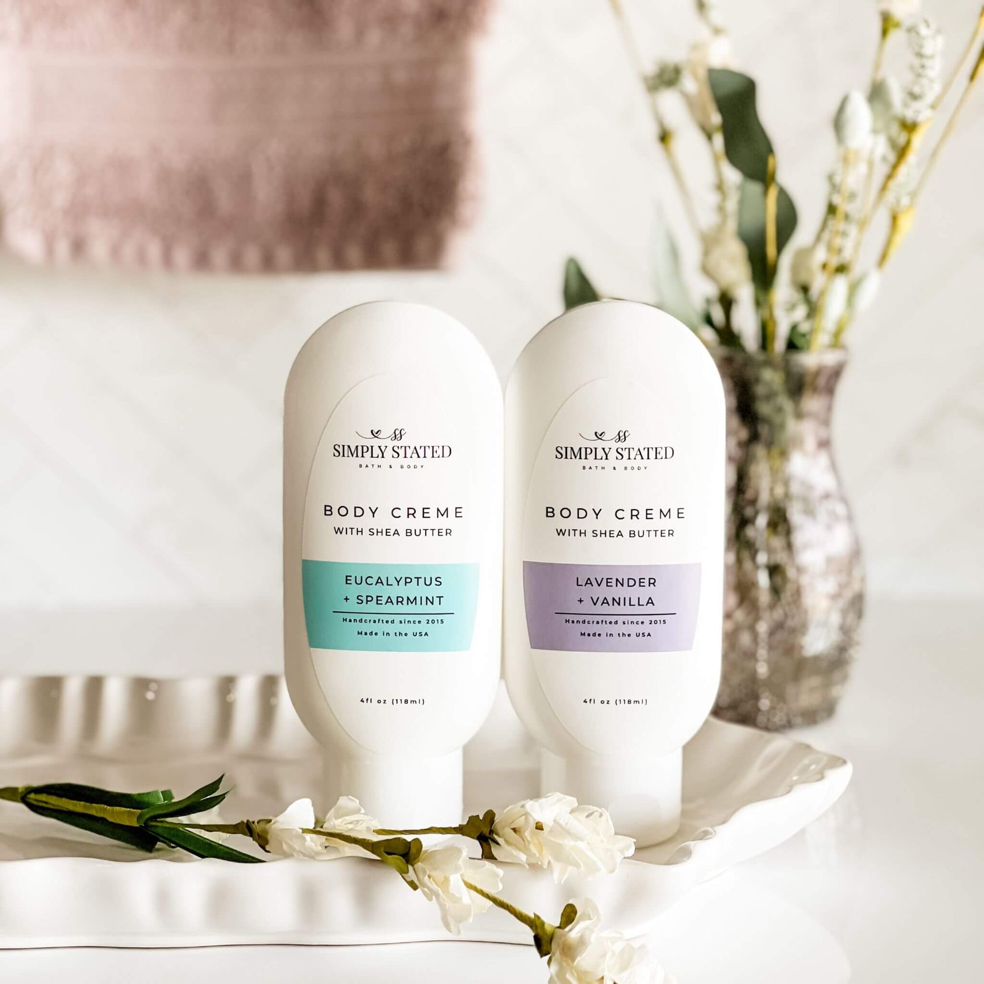 Two bottles of Simply Stated Bath & Body Aromatherapy Body Creme in Eucalyptus + Spearmint and Lavender + Vanilla, displayed on a white tray with floral accents. A soft, neutral background enhances the calming aesthetic.