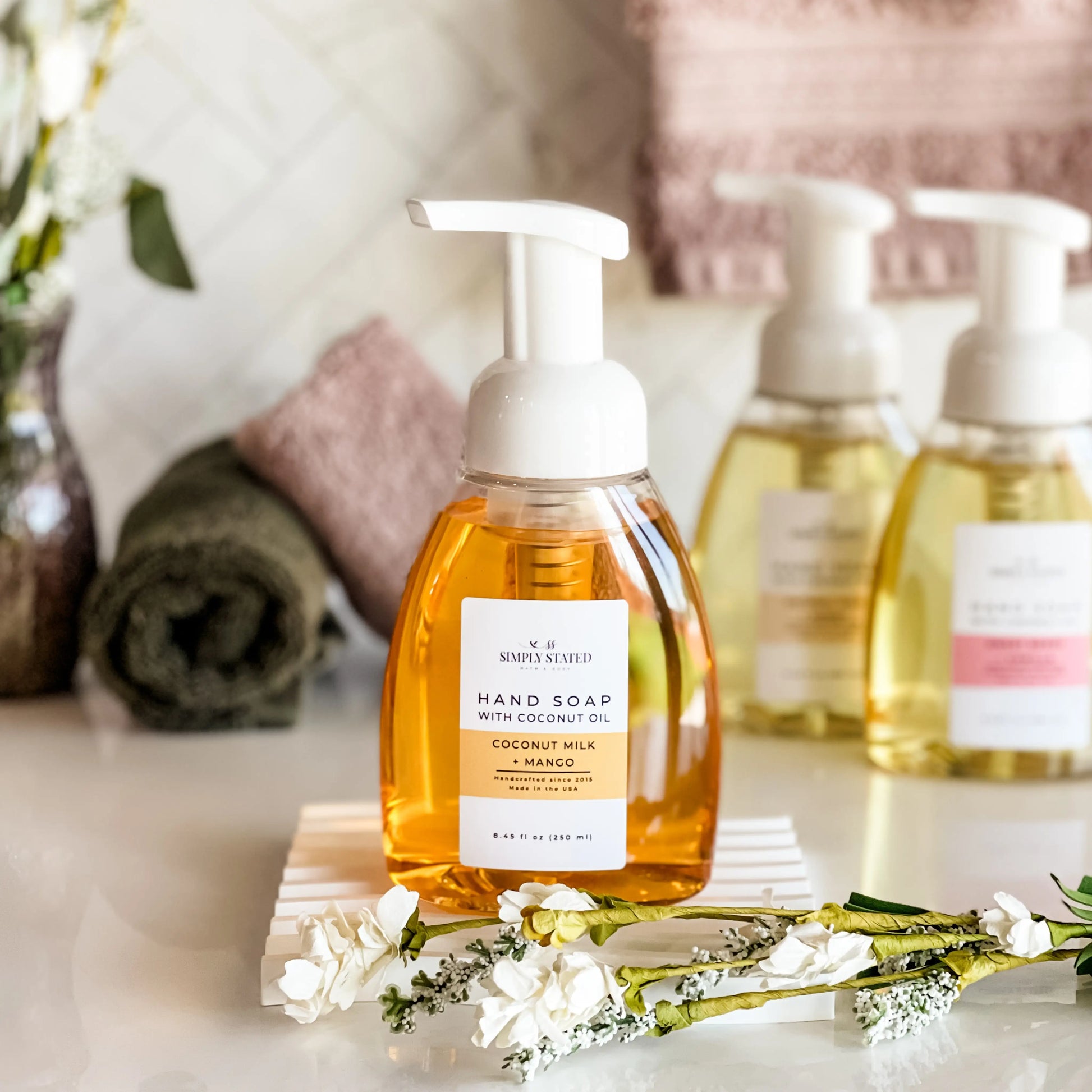 Hand soap bottle with pump dispenser on a wooden block with flowers, surrounded by more soap bottles in the background.
