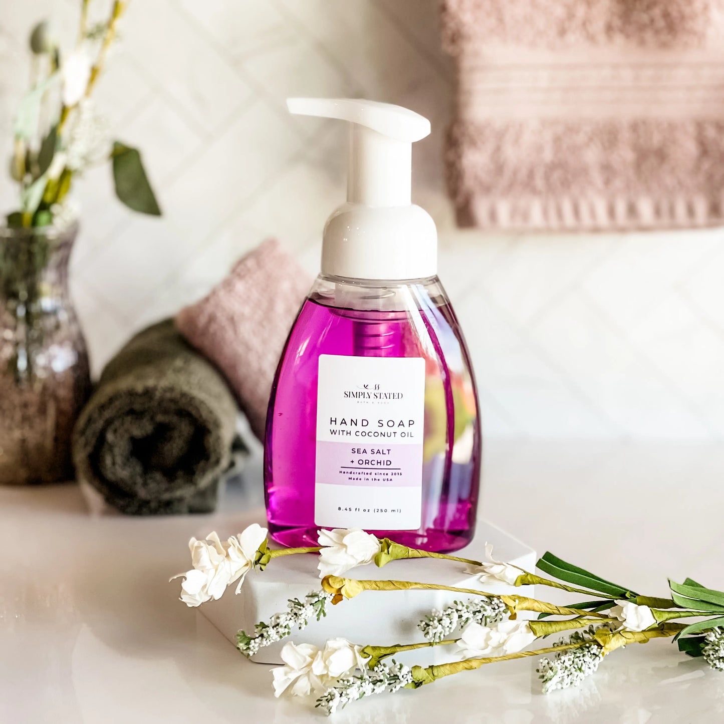 Purple hand soap bottle with pump on a marble surface with flowers and towels in the background