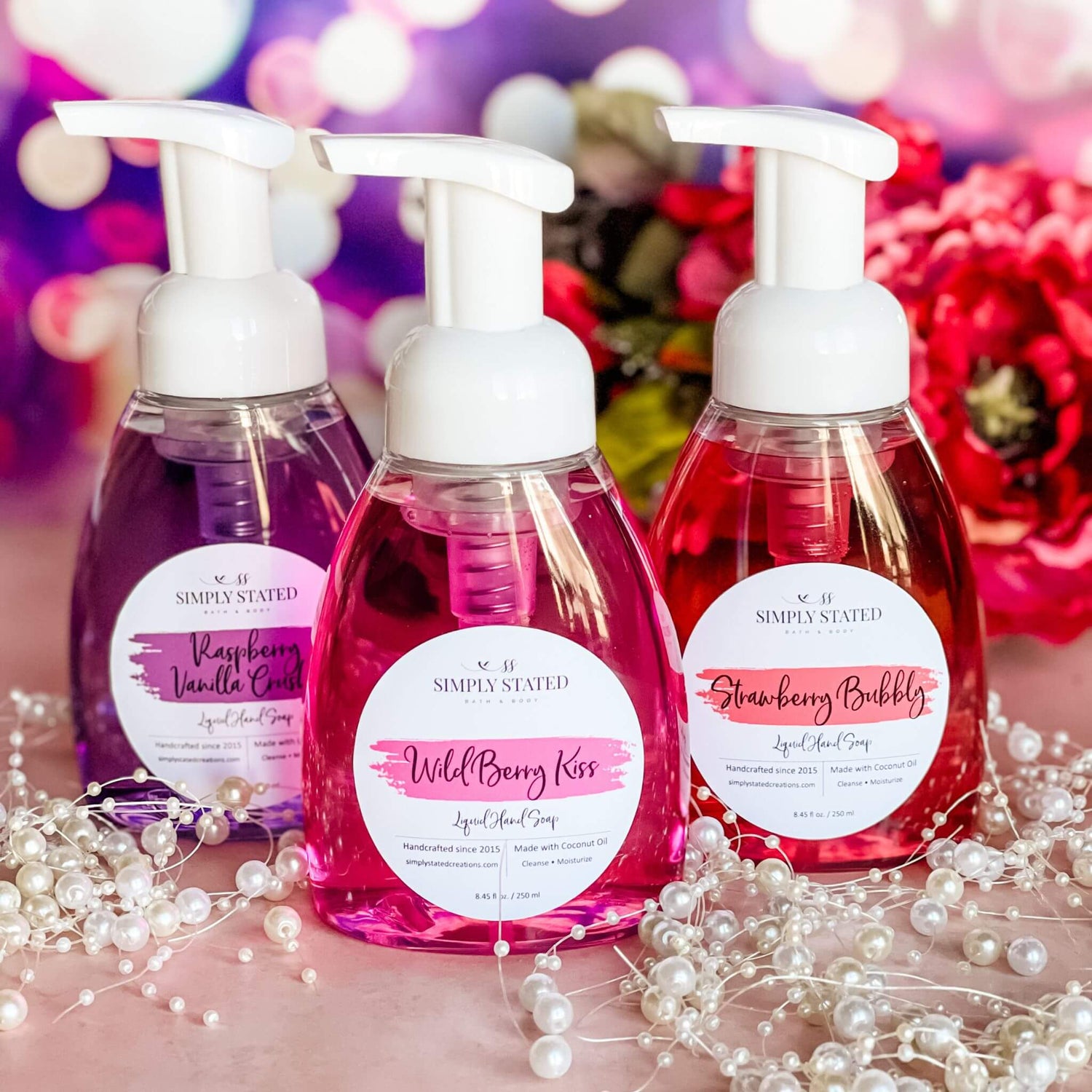 A trio of Sweet Temptations Liquid Hand Soaps—Raspberry Vanilla Crush, Wild Berry Kiss, and Strawberry Bubbly—arranged with pearl accents and vibrant floral decor on a blush pink background.