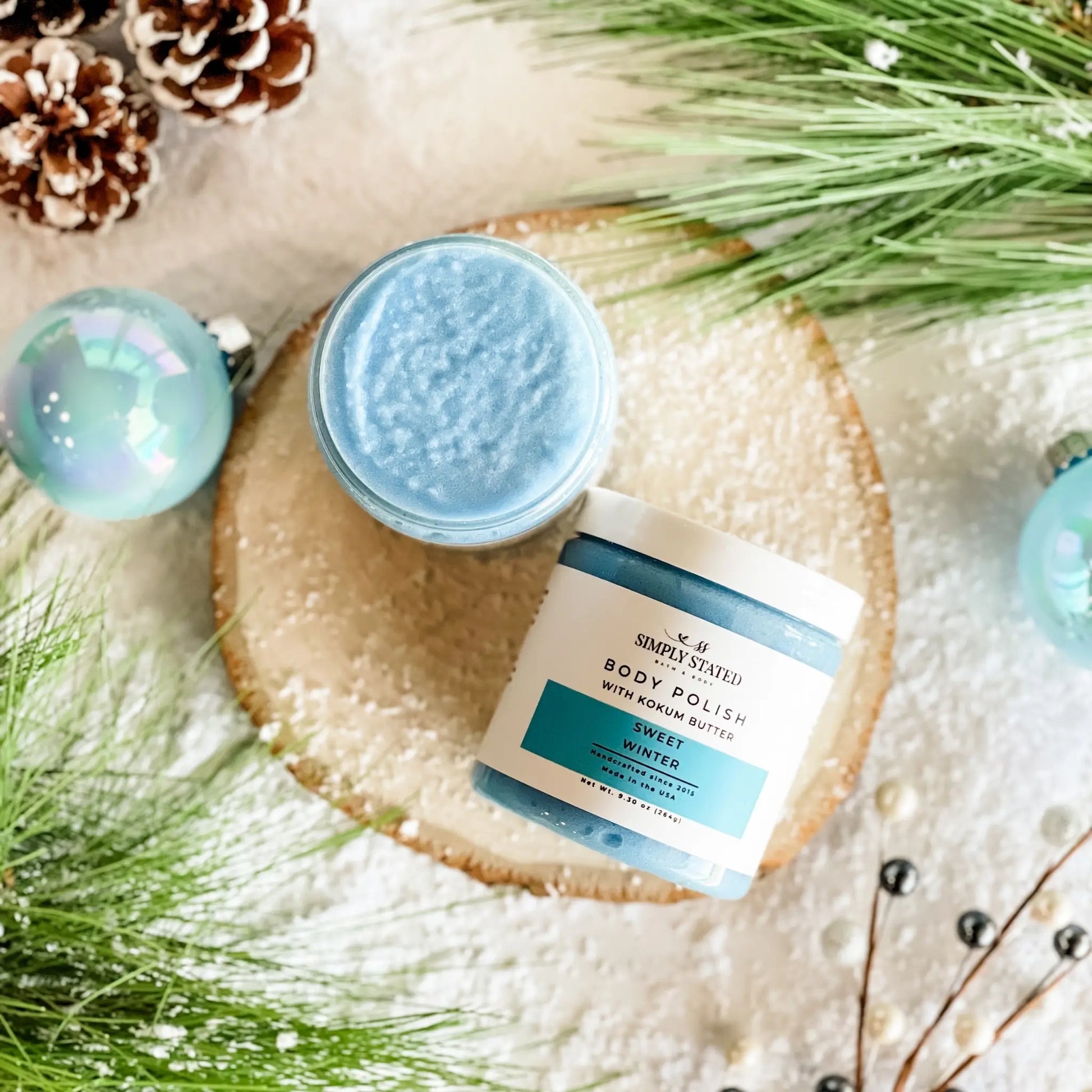 Two jars of body polish on a decorative surface with greenery and ornaments.