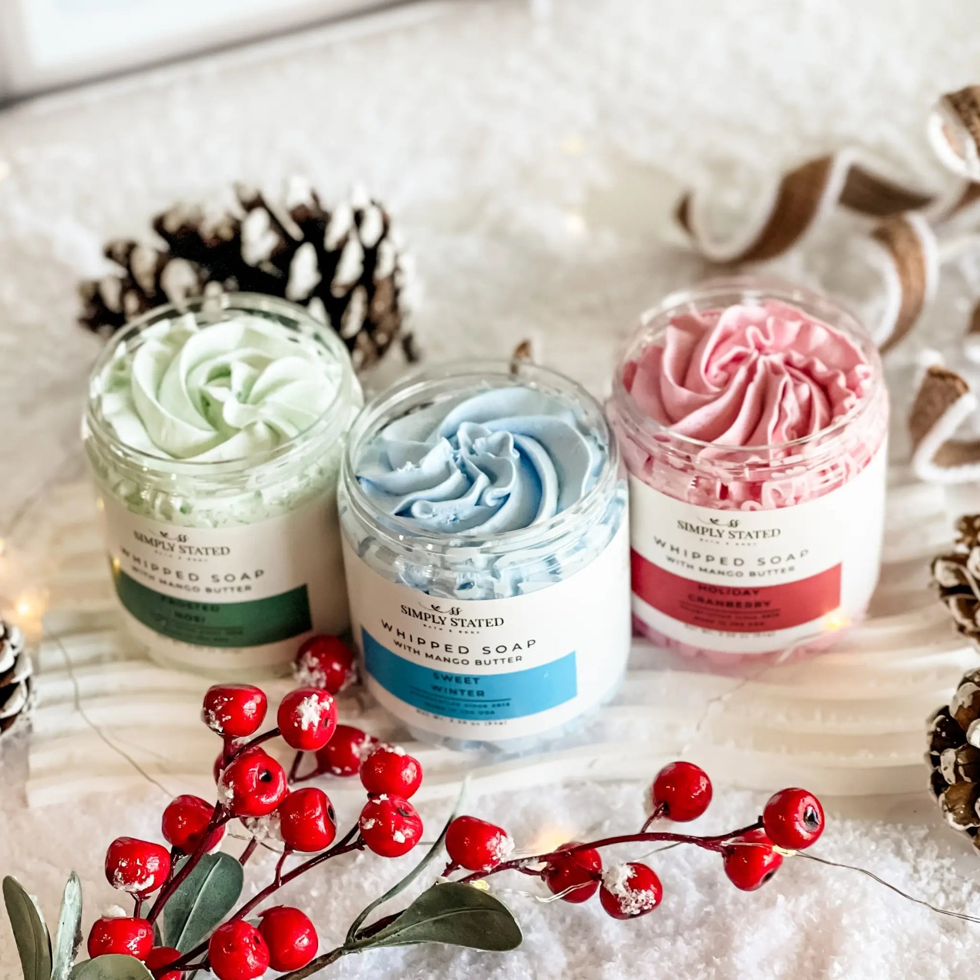 Three jars of whipped soap with decorative berries and pine cones on a textured surface.